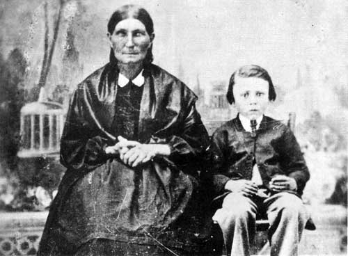 Old photo of a aged woman wearing a black dress, black shawl, braids, sitting beside a child about 10 years old.